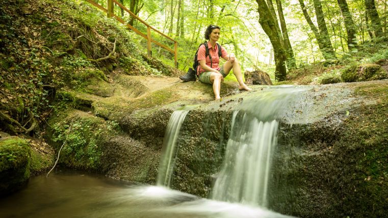 Eine Frau sitzt auf einem Felsen neben einem kleinen Wasserfall im Wald.