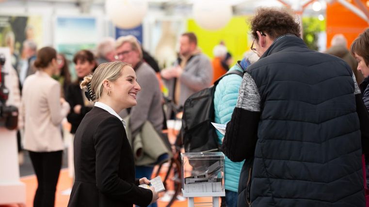 Eine Messe mit vielen Besuchern, die sich unterhalten. Eine lächelnde Frau im Vordergrund spricht mit einem Mann.