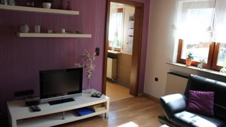 Ein gemütliches Wohnzimmer mit lila Wänden und einem schwarzen Sofa. Es gibt einen Fernseher und einen kleinen Tisch, sowie Fenster mit Pflanzen.