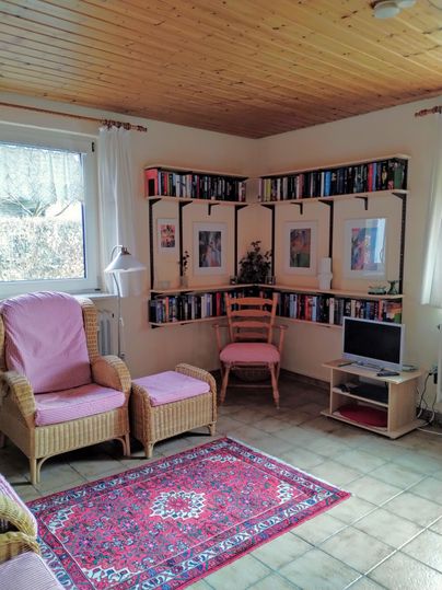 Ein gemütlicher Raum mit einem Bücherregal an der Wand. Es gibt bequeme Sessel, einen kleinen Tisch und einen Teppich.