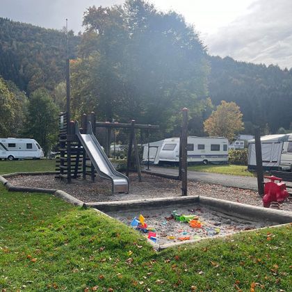 Ein Spielplatz mit einer Rutsche und einer Sandkiste. Im Hintergrund sind Wohnmobile und Bäume zu sehen.