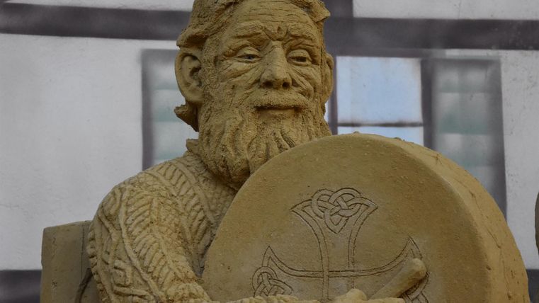 Eine detailreiche Sandskulptur eines bärtigen Mannes, der ein Schild hält. Die Figur zeigt kunstvolle Muster und ist gut gestaltet.