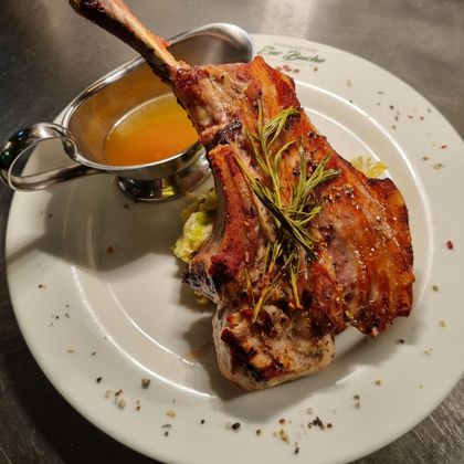 Ein saftiges Stück Fleisch mit einem Knochen, garniert mit frischen Kräutern. Dazu gibt es eine kleine Schüssel mit Sauce auf einem weißen Teller.