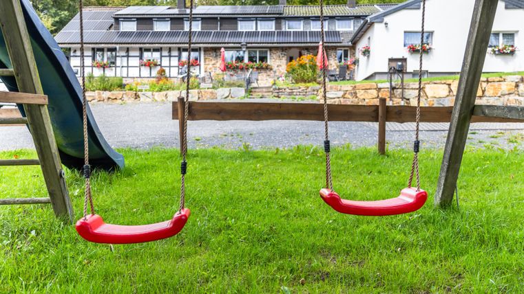 Eine Schaukel im Garten mit grünem Gras im Vordergrund. Im Hintergrund ist ein Gebäude mit Blumenfenstern zu sehen.