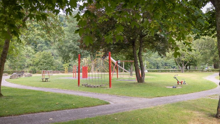 Ein Spielplatz mit roten Spielgeräten steht in einem grünen Park. Der Bereich ist von Bäumen umgeben und bietet eine einladende Atmosphäre.