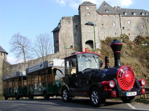 Ein kleiner Zug steht vor einer Burg. Die Umgebung ist grün mit Bäumen und historischem Steinbau.