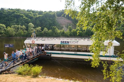 Ein Boot liegt an einer Anlegestelle am Wasser. Menschen steigen ein, während die grüne Landschaft im Hintergrund sichtbar ist.