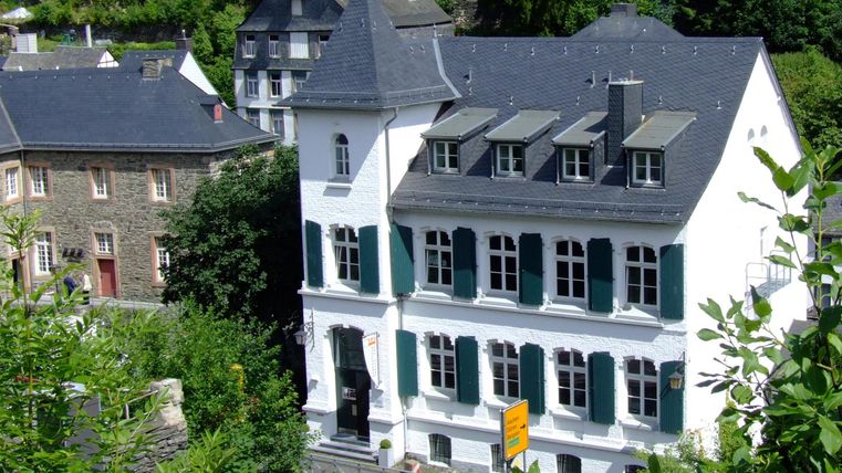 Ein charmantes, weißes Gebäude mit grünen Fensterläden. Umgeben von Bäumen und weiteren historischen Gebäuden.