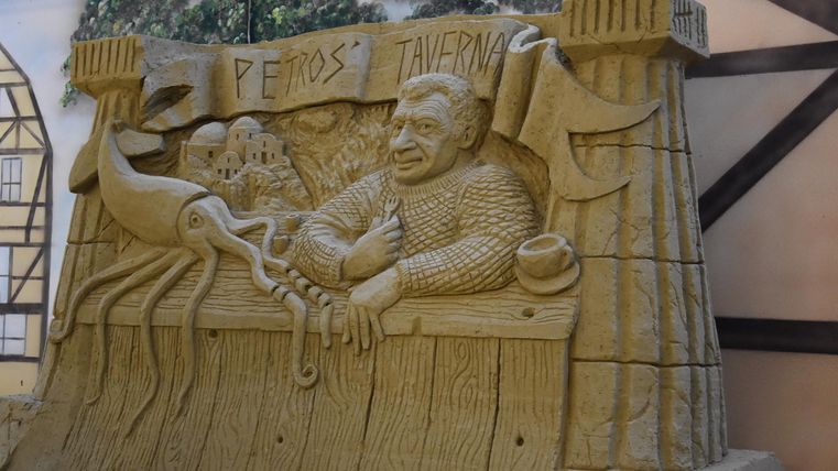 Eine große Sandskulptur mit einer Figur, die an einen Ausschank erinnert. Sie zeigt einen Mann, der freundlich lächelt und von den Meeresfrüchten umgeben ist.