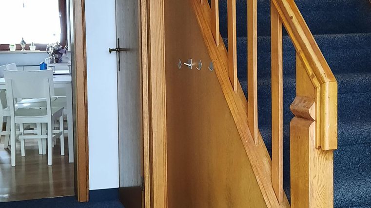 Ein heller Flur mit einer Holztreppe und einem teppichbedeckten Boden. Im Hintergrund ist eine offene Tür zu einem Raum sichtbar.