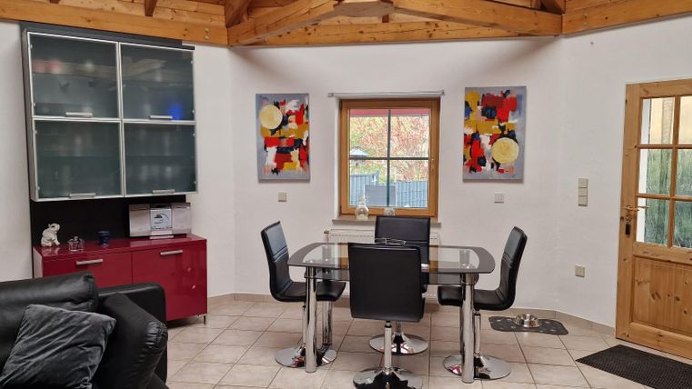 Ein modernes Wohnzimmer mit einem Esstisch und schwarzen Stühlen. An den Wänden hängen bunte Bilder und ein Fenster lässt viel natürliches Licht herein.