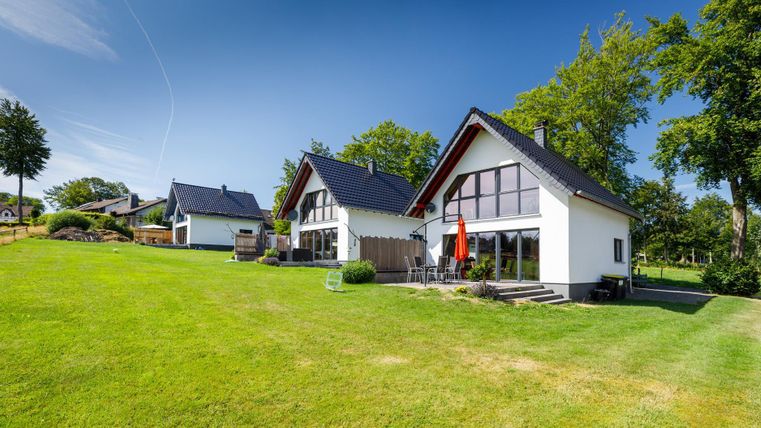 Zwei moderne Häuser stehen auf einer grünen Wiese. Der klare Himmel und die Bäume im Hintergrund verleihen der Szene eine ruhige Atmosphäre.