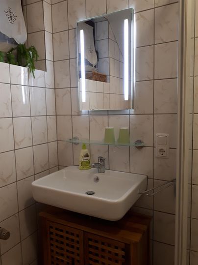 Ein modernes Badezimmer mit einem weißen Waschbecken und einem beleuchteten Spiegel. Die Wände sind gefliest, und es gibt ein Holzregal für Pflegeprodukte.