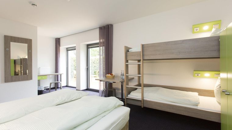 Ein modernes Zimmer mit einem Doppelbett und einem Etagenbett. Große Fenster sorgen für viel Tageslicht und eine freundliche Atmosphäre.