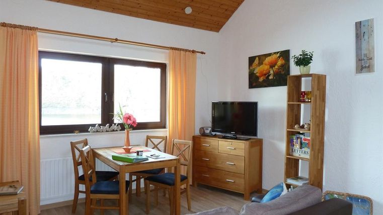 Ein gemütlicher Wohnbereich mit einem Esstisch, Stühlen und einem Fernseher. Große Fenster mit Vorhängen lassen viel Licht hinein und es gibt eine kleine Regal mit Pflanzen und Dekoration.