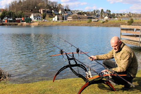 Ein Angler sitzt am Ufer eines Sees und bereitet seine Angelausrüstung vor. Im Hintergrund sind kleine Häuser und Hügel zu sehen.