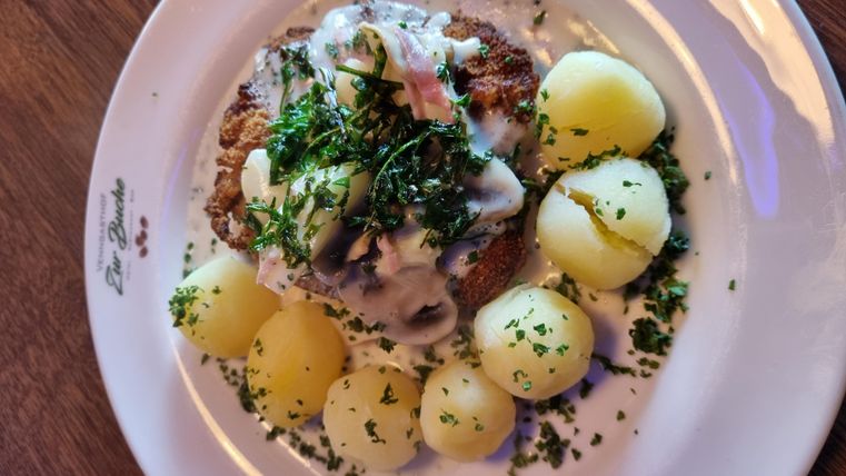 Ein köstliches Gericht mit einem schnitzelartigen Hauptteil, überzogen mit einer weißen Sauce und frischen Kräutern. Dazu gibt es gekochte Kartoffeln, die ordentlich angerichtet sind.
