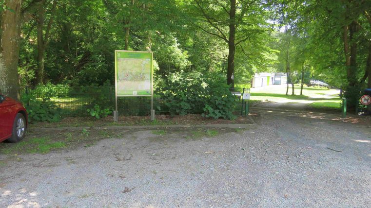 Ein schmaler Schotterweg zwischen Bäumen führt zu einem Parkplatz. Im Hintergrund sind weitere Bäume und ein Schild sichtbar.