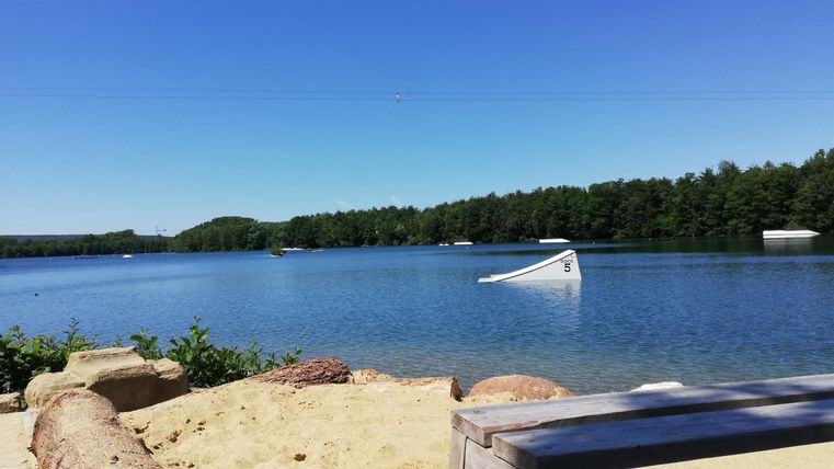 Ein ruhiger See umgeben von Bäumen und sandigem Strand. Der Himmel ist klar und blau, ideal für einen entspannten Tag.