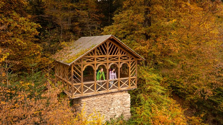 Zwei Personen stehen in einer hölzernen Hütte im Wald, umgeben von herbstlichen Bäumen.