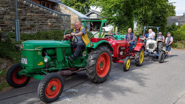 Eine Gruppe von Traktoren fährt eine Straße entlang. Die Fahrzeuge sind bunt und die Umgebung ist ländlich mit Bäumen und Häusern.