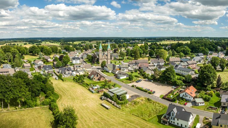 Eine ruhige Landschaft mit einem kleinen Dorf und einer Kirche im Zentrum. Rund um das Dorf befinden sich grüne Felder und Bäume.