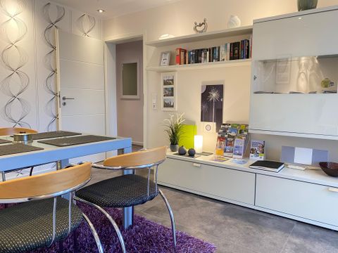 Ein modernes Wohnzimmer mit einem Esstisch und Stühlen. An der Wand befindet sich ein Regal mit Büchern und dekorativen Elementen.