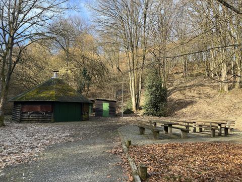 Eine Hütte in einem Waldgebiet mit Laubbäumen. Neben der Hütte steht ein Grillplatz mit Sitzbänken.