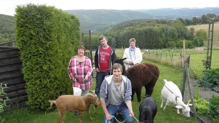 Eine Gruppe von fünf Personen steht in einem Garten mit mehreren Tieren, darunter Ziegen und ein Hund. Im Hintergrund sind Hügel und Bäume zu sehen.