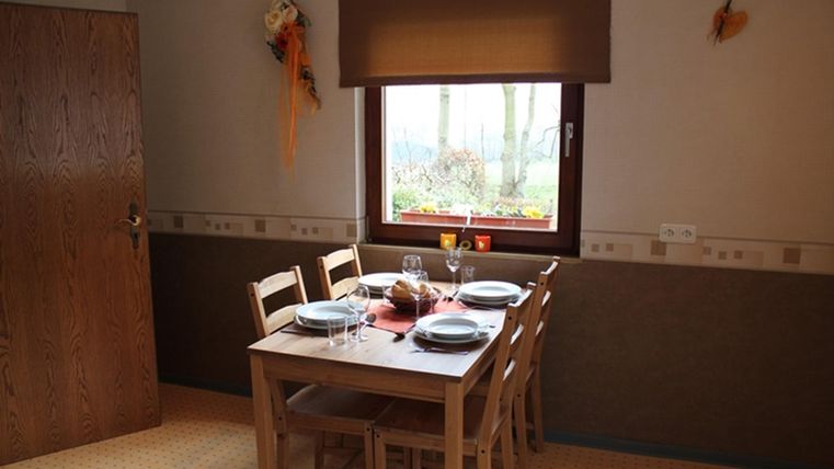 Ein einfacher Essbereich mit einem Holztisch und Stühlen. Am Tisch sind gedeckte Teller und Gläser platziert, und ein Fenster lässt Licht herein.