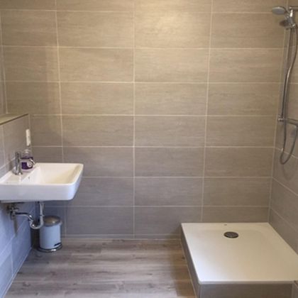 Ein modernes Badezimmer mit grauen Fliesen und einer ebenerdigen Dusche. Das Waschbecken und ein Spiegel sind ebenfalls vorhanden.