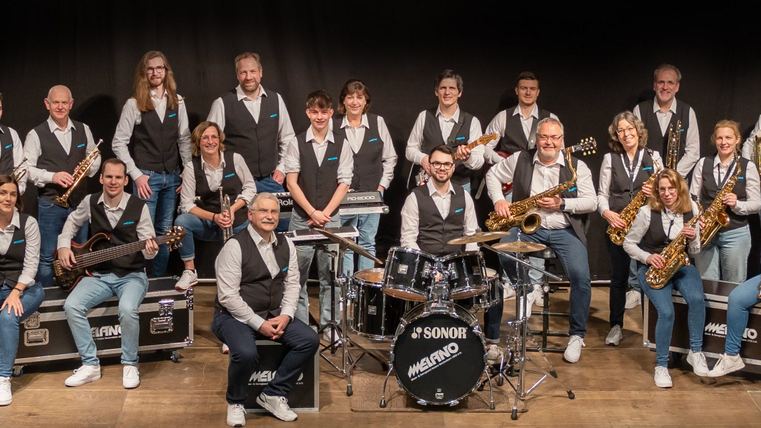 Eine große Musikgruppe steht auf einer Bühne. Sie tragen einheitliche Kleidung und halten verschiedene Musikinstrumente.