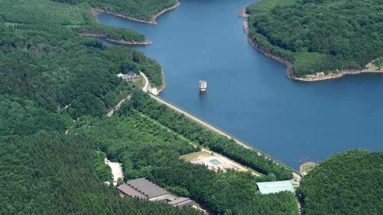 Ein malerischer See umgeben von grünen Wäldern. In der Mitte ist ein kleiner Bootsanleger sichtbar.