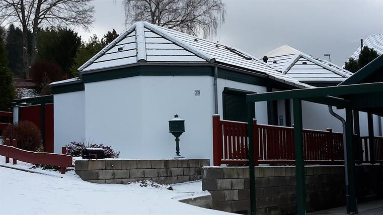 Ein modernes, weißes Haus mit einem flachen Dach, umgeben von Schnee. Im Vordergrund ist ein roter Zaun sichtbar, der das Grundstück umgibt.
