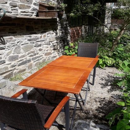 Ein Holztisch mit zwei Stühlen steht in einem begrünten Außenbereich. Im Hintergrund sind eine Steinmauer und Pflanzen zu sehen.