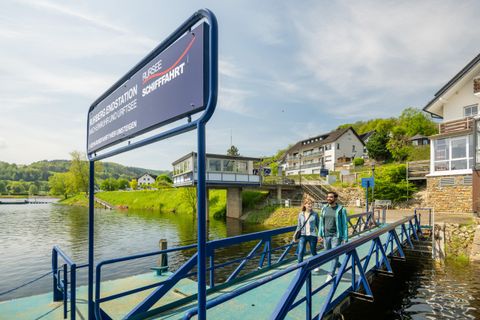 Eine entspannte Umgebung am Wasser mit einem Steg und einem Informationsschild zur Schifffahrt. Zwei Personen spazieren am Ufer entlang und genießen die Aussicht.