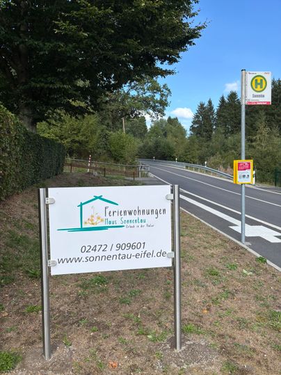 Ein Wegweiser zu Ferienwohnungen namens "Haus Sonnentau" steht an einer Straße. Daneben befindet sich eine Bushaltestelle in einer ländlichen Umgebung.