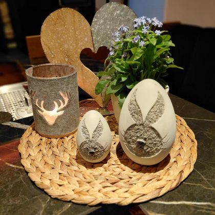 Eine dekorative Anordnung von Frühlingsaccessoires auf einem Tisch. Es sind zwei Eier mit Hasenmotiven, ein kleiner Behälter mit einem Hirschmotiv und eine blühende Pflanze in einem Topf zu sehen.