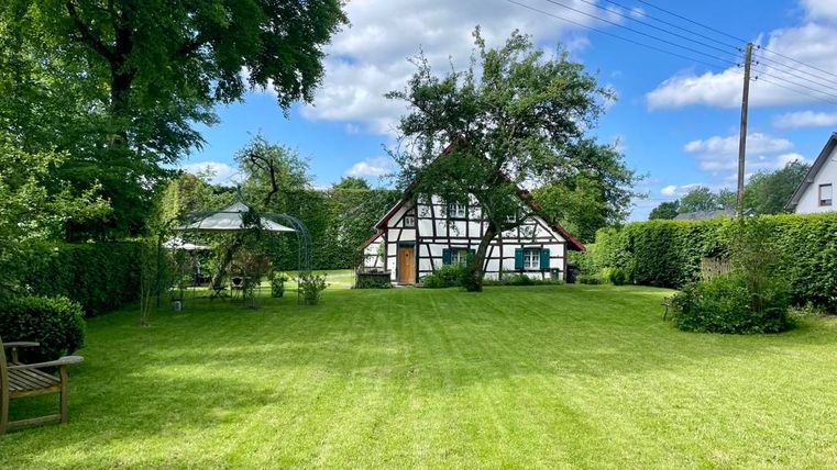 Ein schöner Garten mit grünem Rasen und einem traditionellen Fachwerkhaus. Im Hintergrund gibt es viele Bäume und einen klaren blauen Himmel.