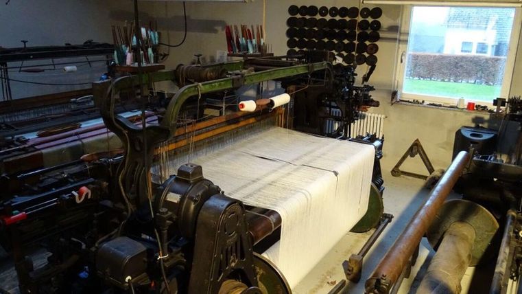 Eine alte Webmaschine in einem werkstattähnlichen Raum. Der Tisch ist mit einem großen Stück gewebtem Stoff bedeckt und bietet einen Einblick in die Textilproduktion.