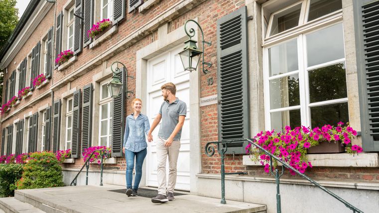 Ein charmantes Gebäude mit Fenstern, die mit grünen Fensterläden und bunten Blumen geschmückt sind. Zwei Personen stehen auf der Treppe und lächeln.