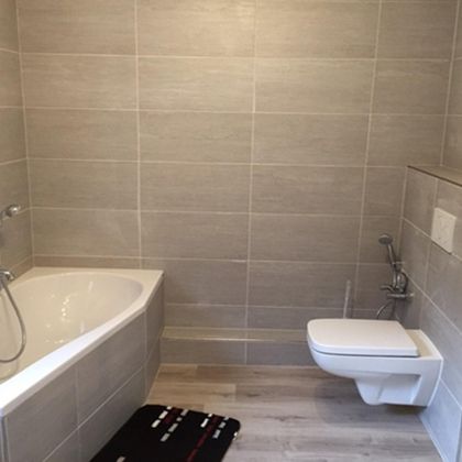Ein modernes Badezimmer mit einer Badewanne und einem WC. Die Wände sind mit grauen Fliesen gekleidet und der Boden ist aus Holz.