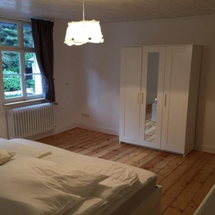 Ein einfaches Schlafzimmer mit einem großen Bett und Holzfußboden. Ein heller Kleiderschrank und ein Fenster mit Vorhänge sorgen für eine angenehme Atmosphäre.
