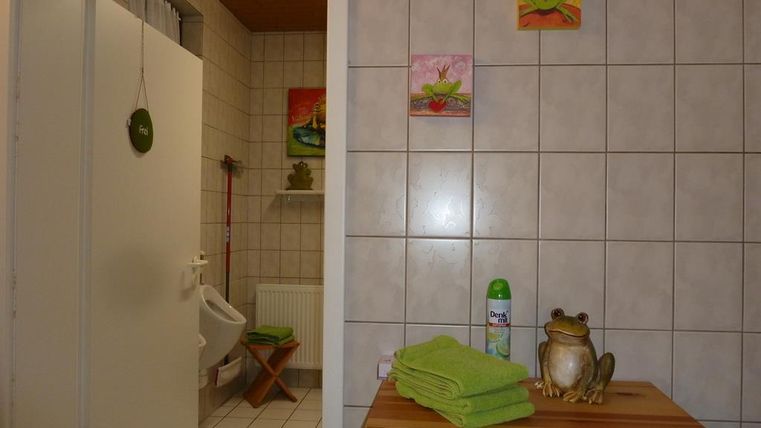 Ein Badezimmer mit weißen Fliesen und einem hellen Bereich. Es gibt grüne Handtücher, eine Fruchtflasche und dekorative Bilder an der Wand.