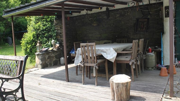 Ein gemütlicher überdachter Essbereich im Freien mit einem Tisch und Stühlen. Umgeben von grünen Pflanzen und einem Holzstamm als Hocker.