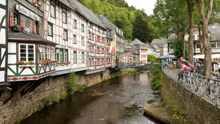 Fachwerkhäuser in Monschau an einem Fluss mit Brücke und Spaziergängern.