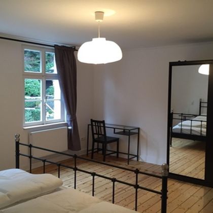 Ein einfaches Schlafzimmer mit einem Doppelbett, einem Schreibtisch und einem großen Spiegel. Helle Wände und Holzfußboden sorgen für eine warme Atmosphäre.