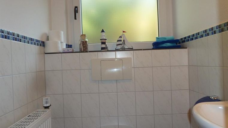 Ein einfaches Badezimmer mit weißen Fliesen und einem Fenster mit Milchglas. Auf der Ablage befinden sich Toilettenpapier, Handtücher und eine dekorative Figur eines Schiffs.