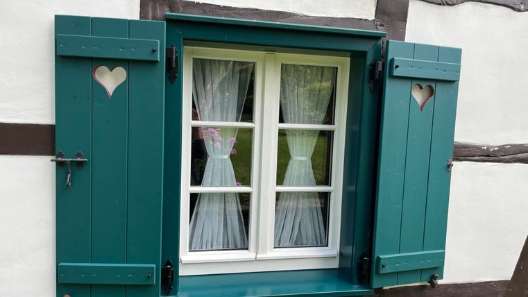 Ein charmantes Fenster mit grünen Fensterläden und weißen Vorhängen. Die Fensterläden haben herzförmige Ausschnitte und verleihen dem Gebäude einen gemütlichen Charakter.