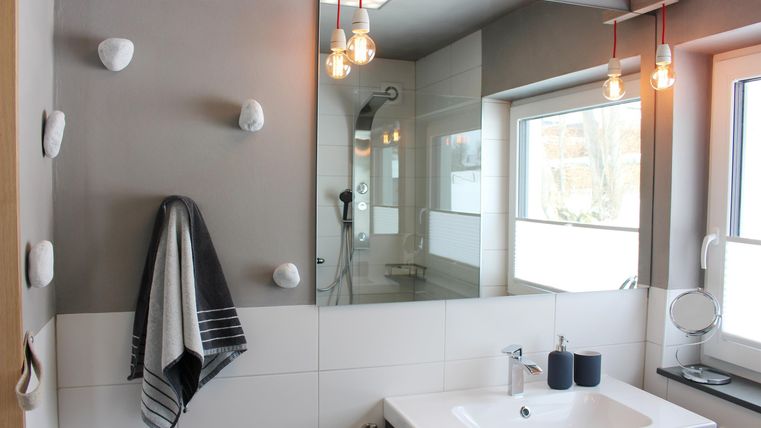 Ein modernes Badezimmer mit einem weißen Waschbecken, einem großen Spiegel und stilvollen Lampen. Die Wände sind hell und der Raum ist gut beleuchtet mit natürlichem Licht.